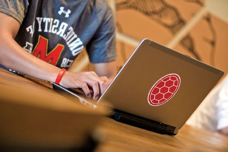 A picture of a male student using his laptop on a table in Starbucks with another student in the background, shell sticker on the laptop